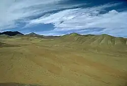À l'avant-plan s'étend une surface déserte présentant différentes couleurs automnales. À l'arrière-plan apparaît des montagnes survolées par des nuages.