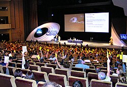 Photographie d'un amphithéâtre où de nombreuses personnes votent en levant la main avec un carton coloré.