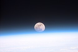 La Lune est visible au-dessus de la Terre et son atmosphère bleutée, formant un disque légèrement aplati.