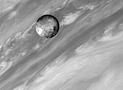 Photographie d'un corps planétaire couvert de nombreuses taches sombres devant les nuages brillants et sombres de Jupiter.
