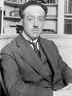 Photographie en noir et blanc d'un homme assis dans une bibliothèque, portant un costume veste-gilet-cravate, mains croisées, moustache courte, front dégarni, l'air impassible.